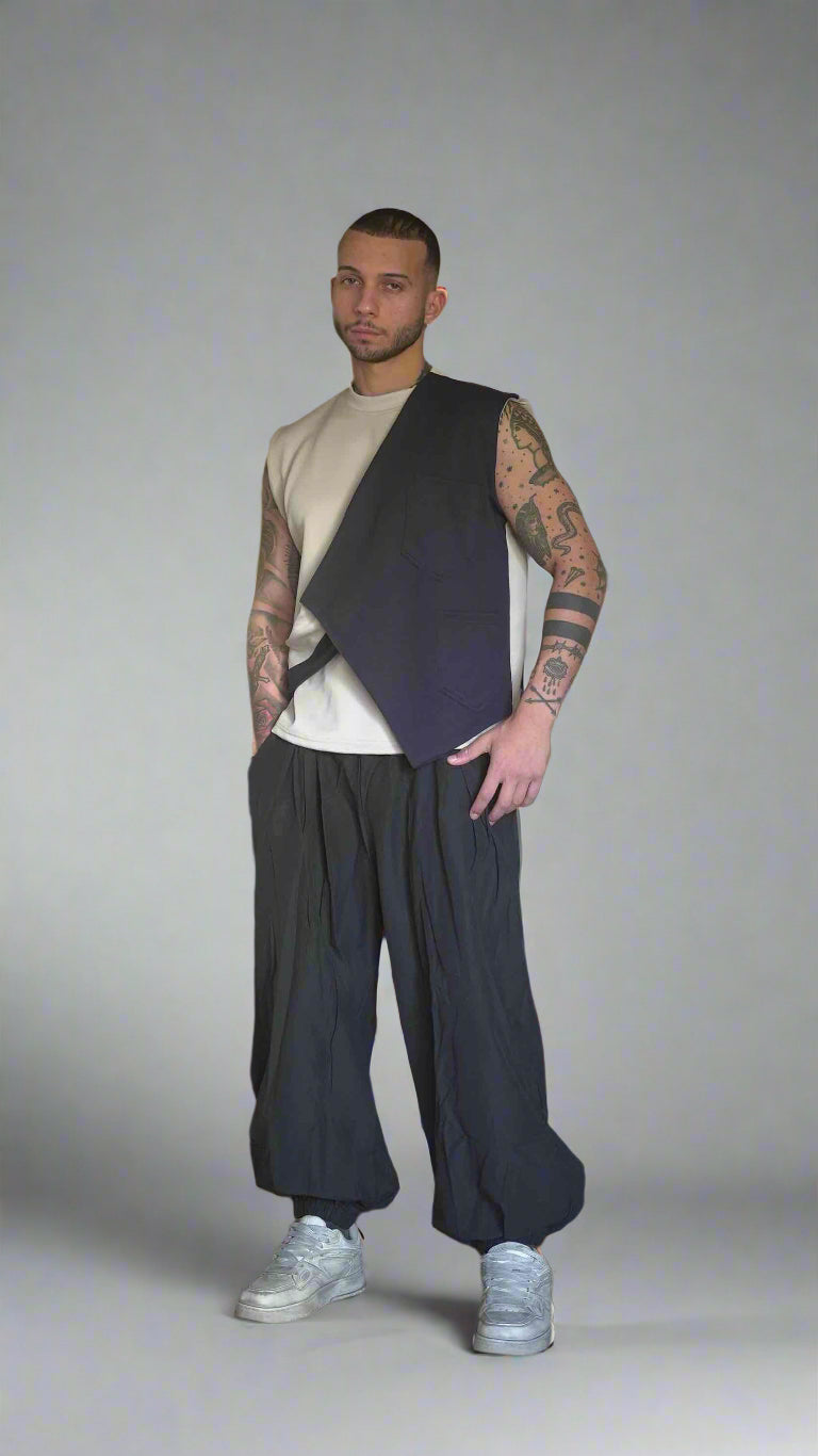 Man with tattoos wearing a vest and pants in a room with furniture and decor.